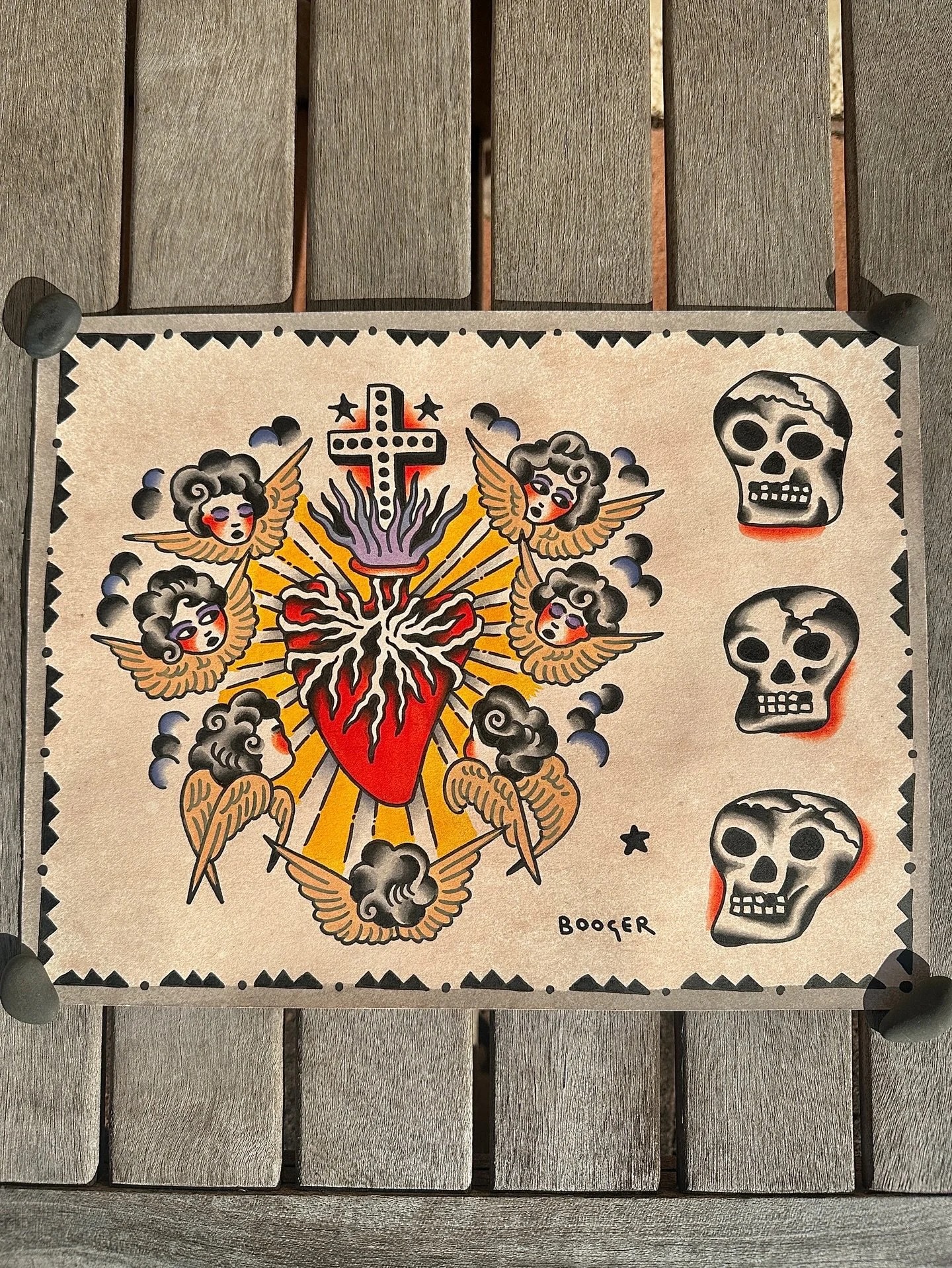 A tattoo design featuring a heart with flames, surrounded by six angelic cherubs with wings, and a cross emitting purple smoke. To the right, there are three skull illustrations with slight variations in appearance. The artist's signature, Booger, is