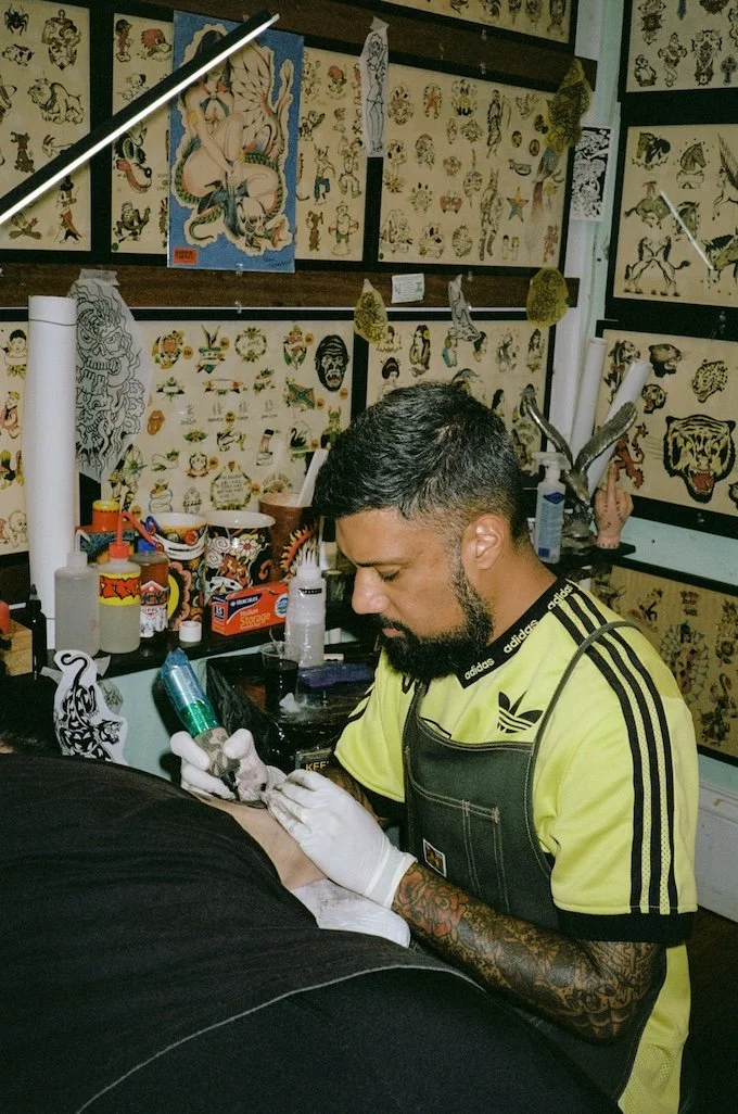 Tattoo artist in yellow Adidas shirt and apron tattooing a person's arm in a tattoo studio decorated with traditional tattoo flash art.