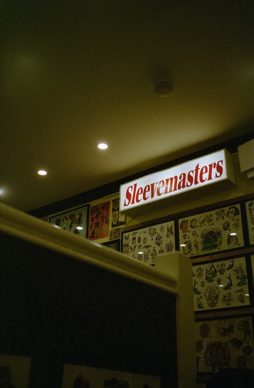 A sign with the word 'Sleepyemasters' in red letters on a white background, inside a room with tattoo sheets on the wall and ceiling lights.