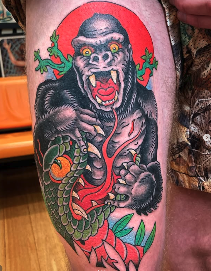 Colorful tattoo of a fierce gorilla with sharp teeth, holding a green snake with an orange fruit, set against a red background with green foliage.