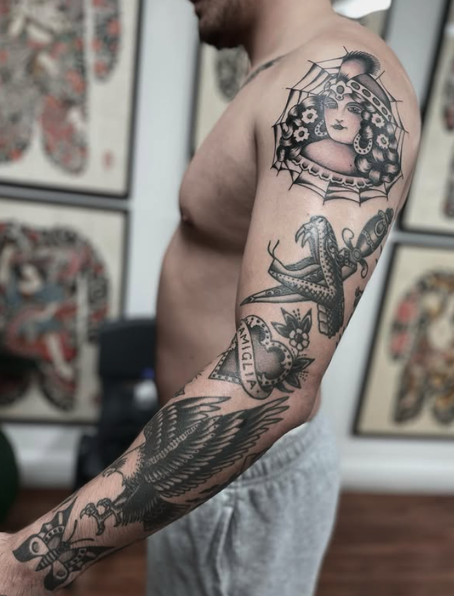 Close-up of a shirtless man with intricate black and gray tattoos on his right arm, chest, and shoulder, featuring a woman inside a parasol, a snake, an eagle, a butterfly, and a dagger, with a background of framed tattoo art on the wall.