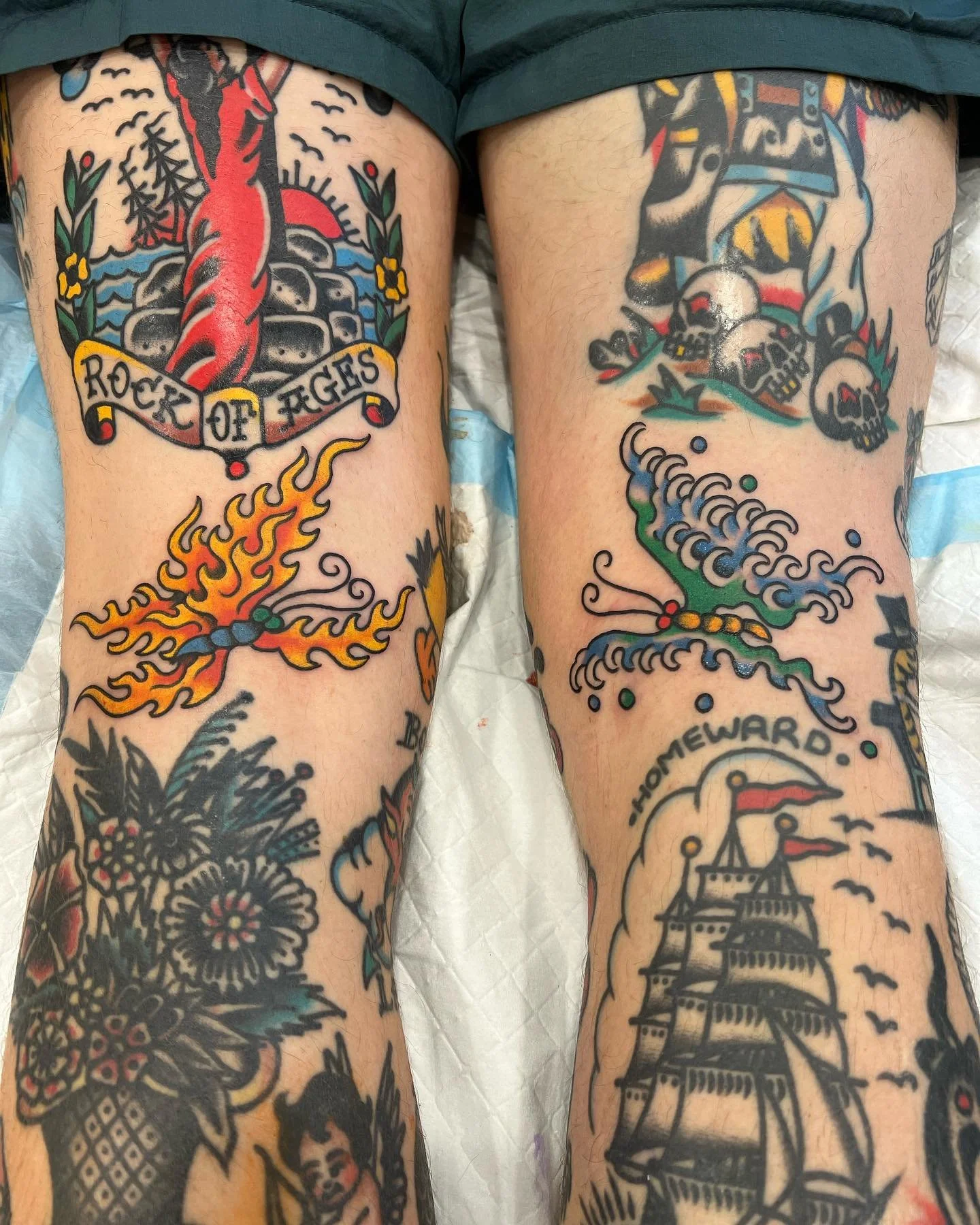 Tattooed legs featuring a flaming butterfly, a traditional ship, skulls with a bandana, flowers, and a banner reading 'Homeward.'
