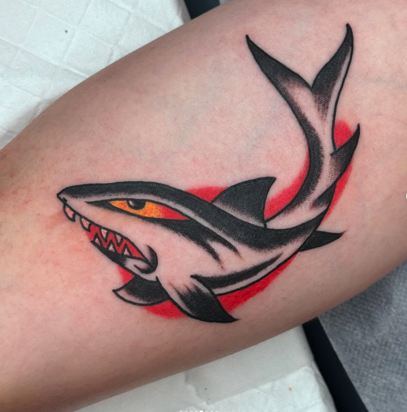 A tattoo of a stylized shark with a menacing expression, sharp teeth, and yellow eye, with a red background on skin.
