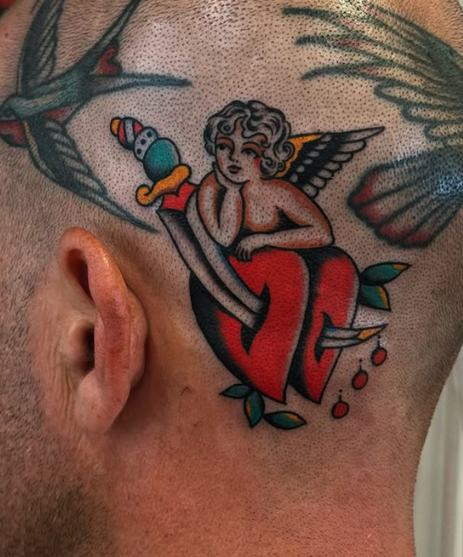 Tattoo of a cherub-like figure with wings, resting on a large red heart with a sword, on the side of a person's head.