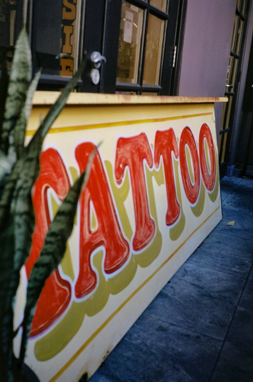 A street sign with the word "Bakery" in red letters, leaning against a wall with a window, partially obscured by plant leaves.