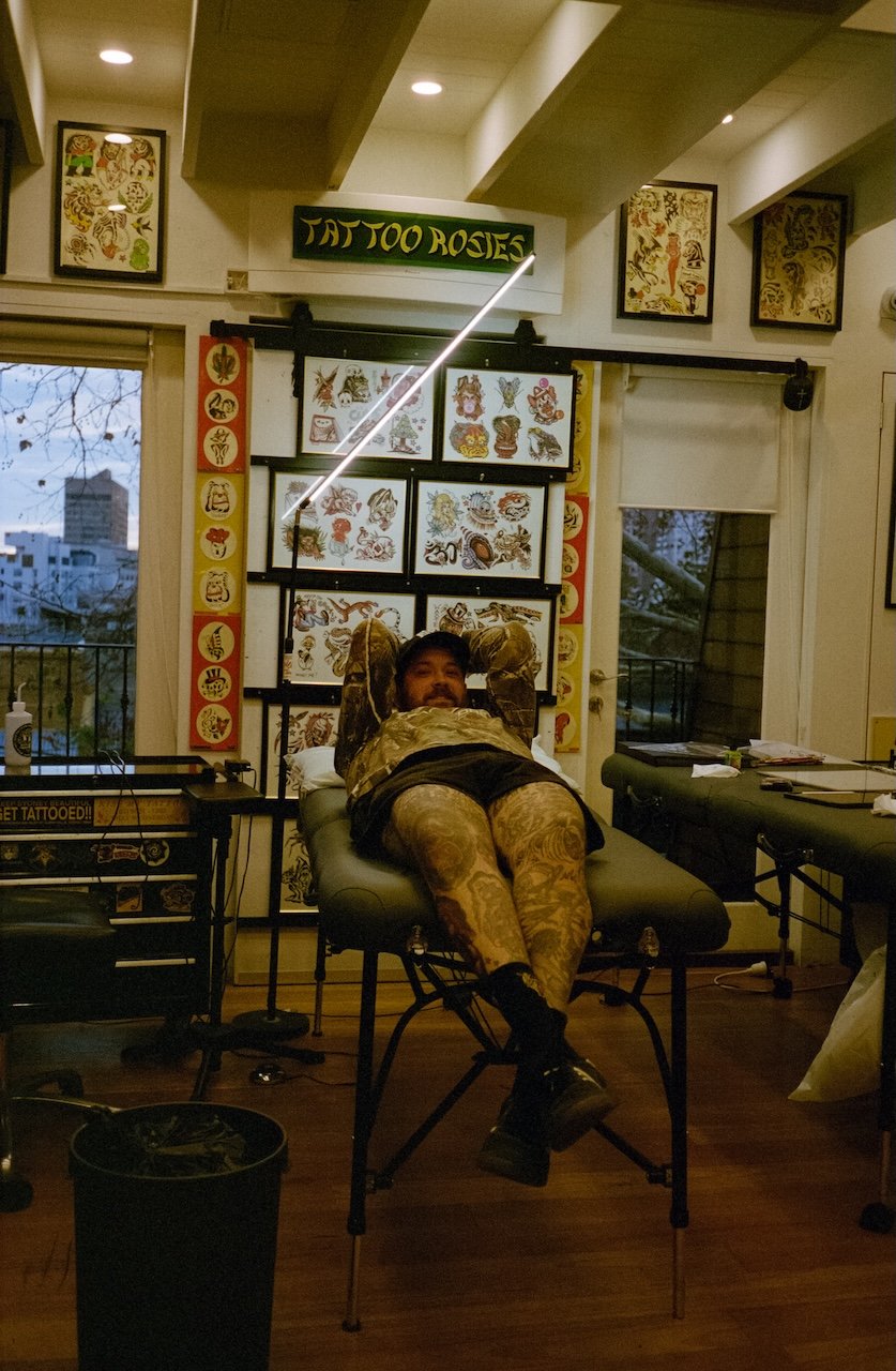 Person lying on a tattoo table inside a tattoo shop, with tattoo designs displayed on the wall behind.