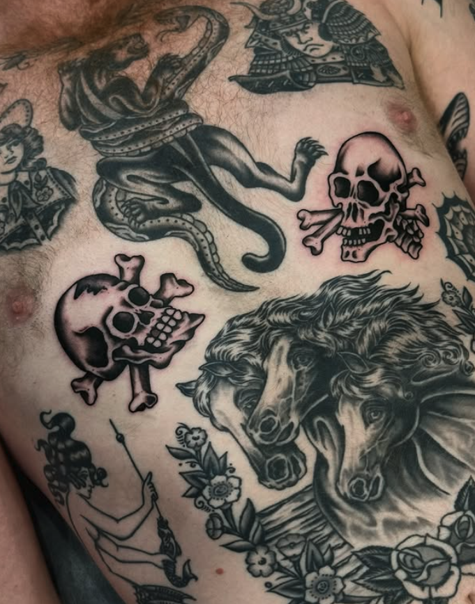 Close-up of a tattooed chest with various designs including two horse heads, skulls, a snake, a knight's helmet, a figure with a spear, and floral elements.