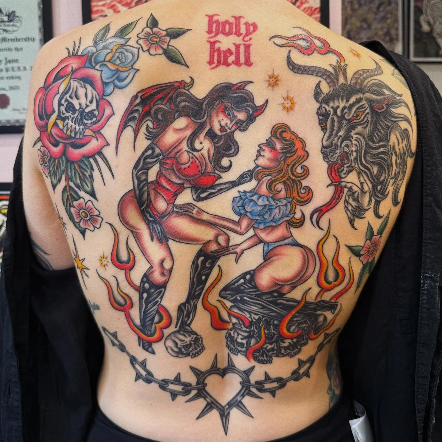 A colorful tattoo on a person's back depicting two women with exaggerated features, surrounded by flames, a lion's head, a skull flower, and the words 'holl y hell' in red stylized lettering at the top.