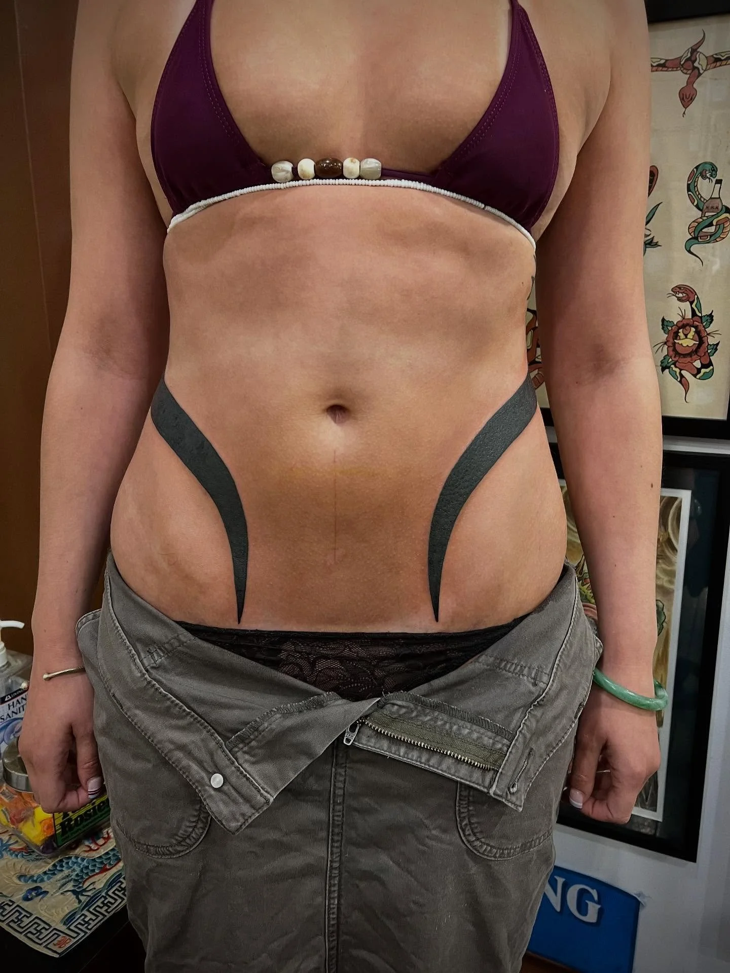 A woman wearing a purple bikini top, black tattoo-like body art on her torso, gray pants that are unzipped, and black lace undergarments, standing indoors.