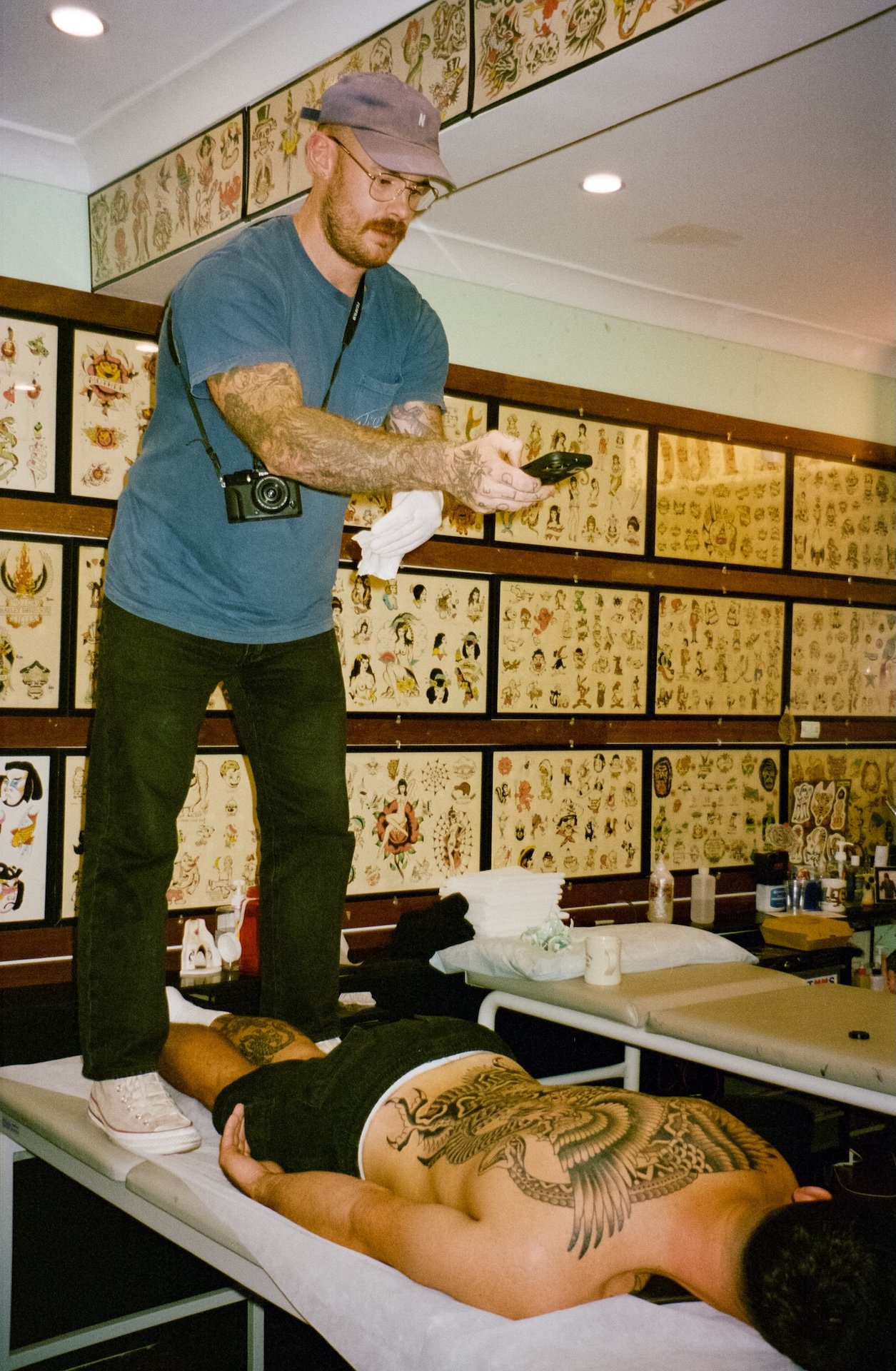 A man standing on a tattooed person's back in a tattoo parlor, with tattoo flash art on the walls.