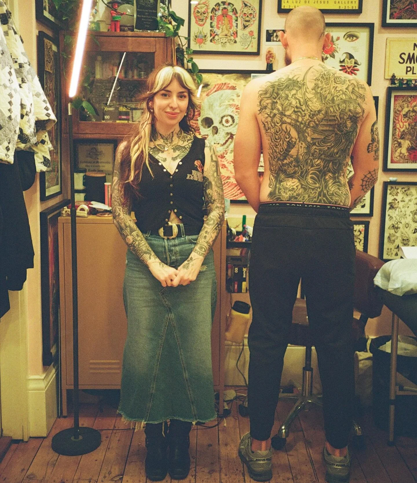A woman with long hair and tattoos on her arms standing in front of a man with tattoos on his back, both in tattoo shop or art studio with framed art and skull paintings on the wall.