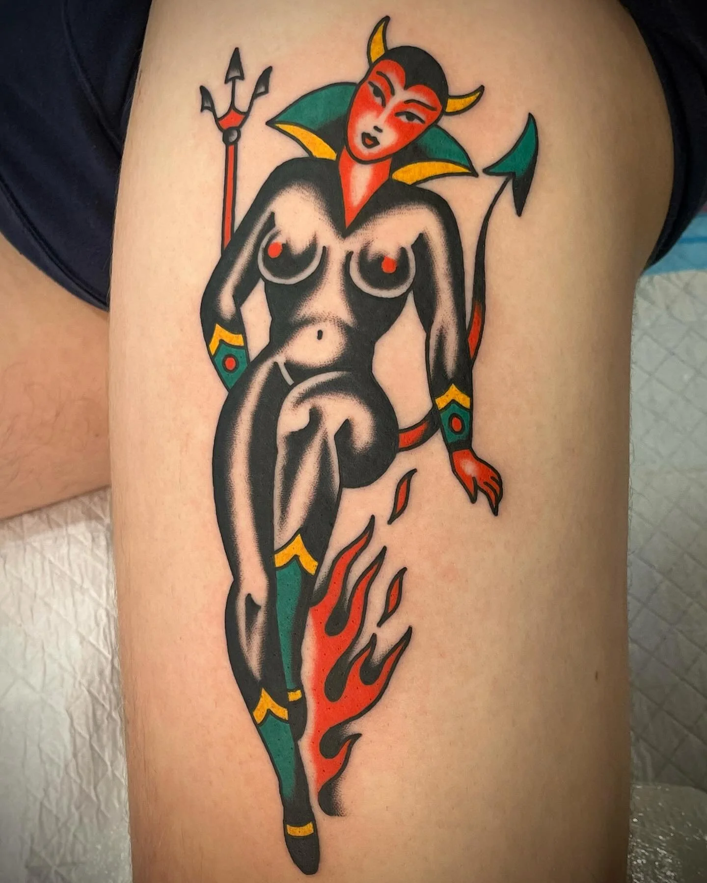 Tattoo of a stylized devil woman with horns, a tail, and a trident, featuring red, black, and green colors, with flames below her.