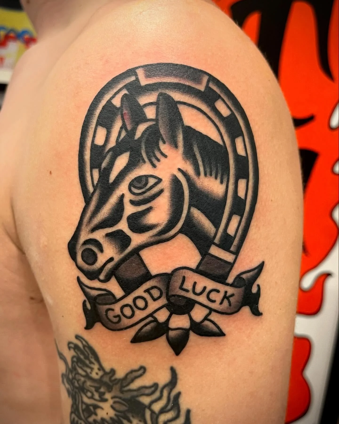Black and grey tattoo of a horse's head within a circular frame, with a ribbon below that reads "Good Luck" and a decorative flower underneath.