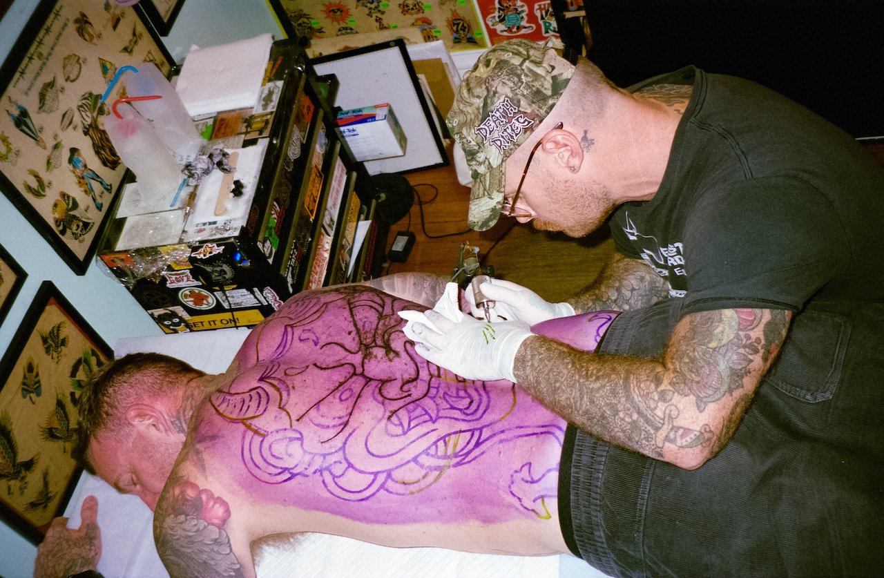 A man receiving tattoo work on his back by a tattoo artist in a tattoo shop filled with tattoo flash artwork.