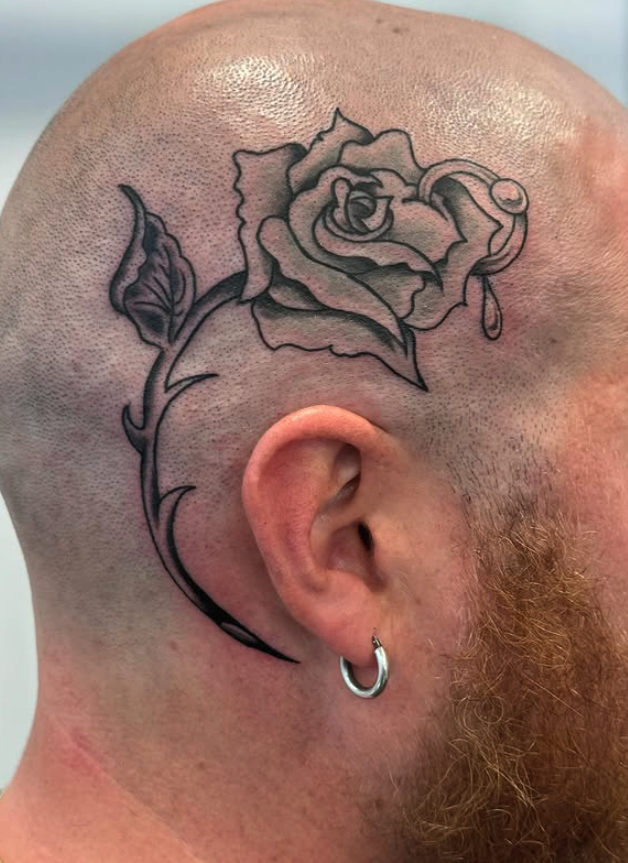 Black outline tattoo of a rose with leaves behind a person's ear on a shaved head, with a small tear drop detail on the rose.