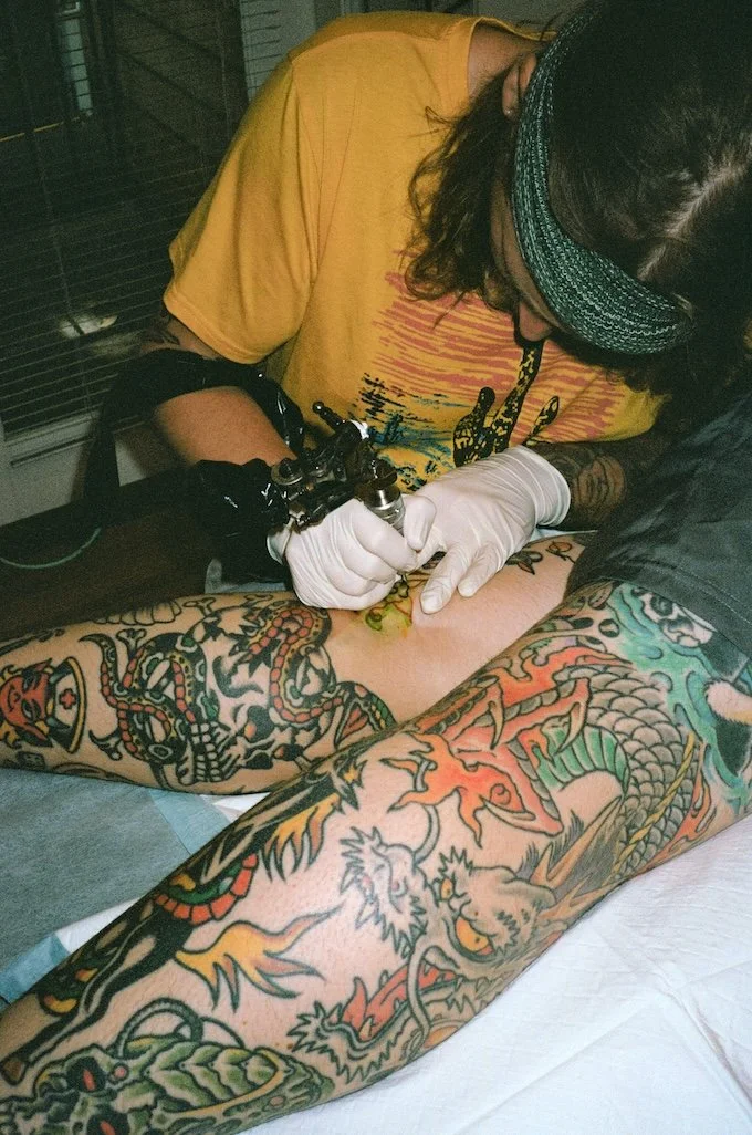 A tattoo artist wearing glasses and a headband is tattooing a large, colorful dragon and tiger design on a person's arm. The artist is using a tattoo machine and wearing white gloves.