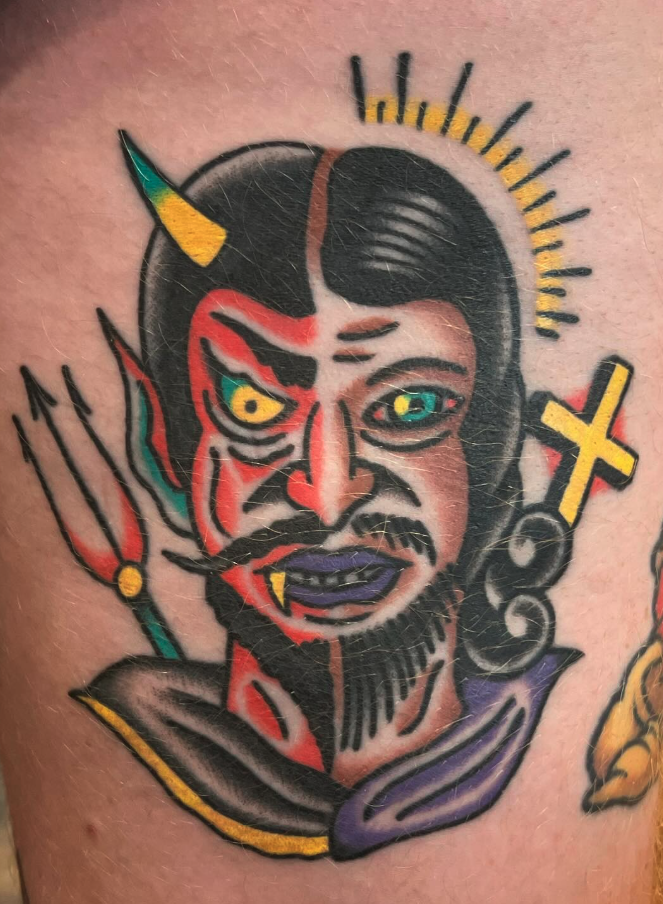 Colorful tattoo of a demonic-looking face with yellow eyes, fangs, and a bristling mustache and beard, surrounded by symbolic elements including a yellow halo above the head, a yellow cross, and a trident. The face has a split design with one side ap