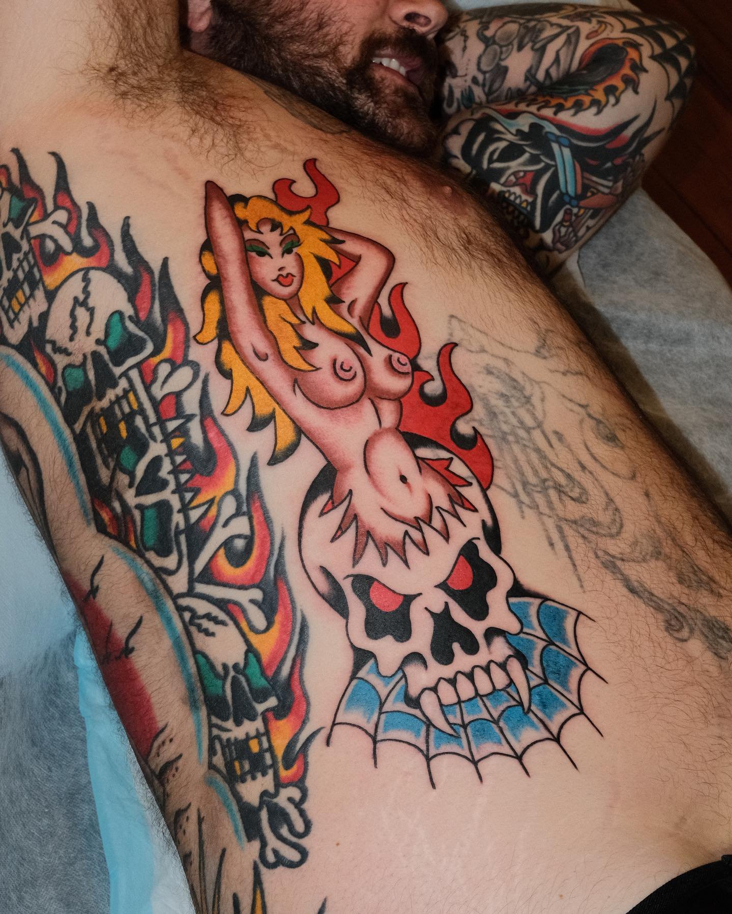 Tattoo of a cartoonish naked woman with long, flowing yellow hair and green eyes, posed with arms behind her head, above a skull with red eyes and a spiderweb pattern on the chin, surrounded by flames and skulls.