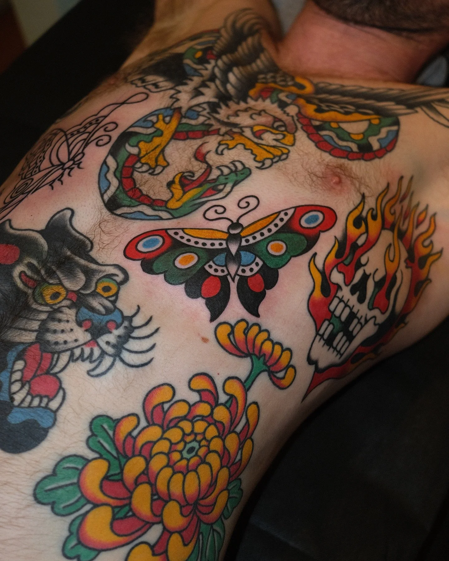 Tattooed chest featuring a colorful dragon, butterfly, skull with flames, lion face, and floral designs.