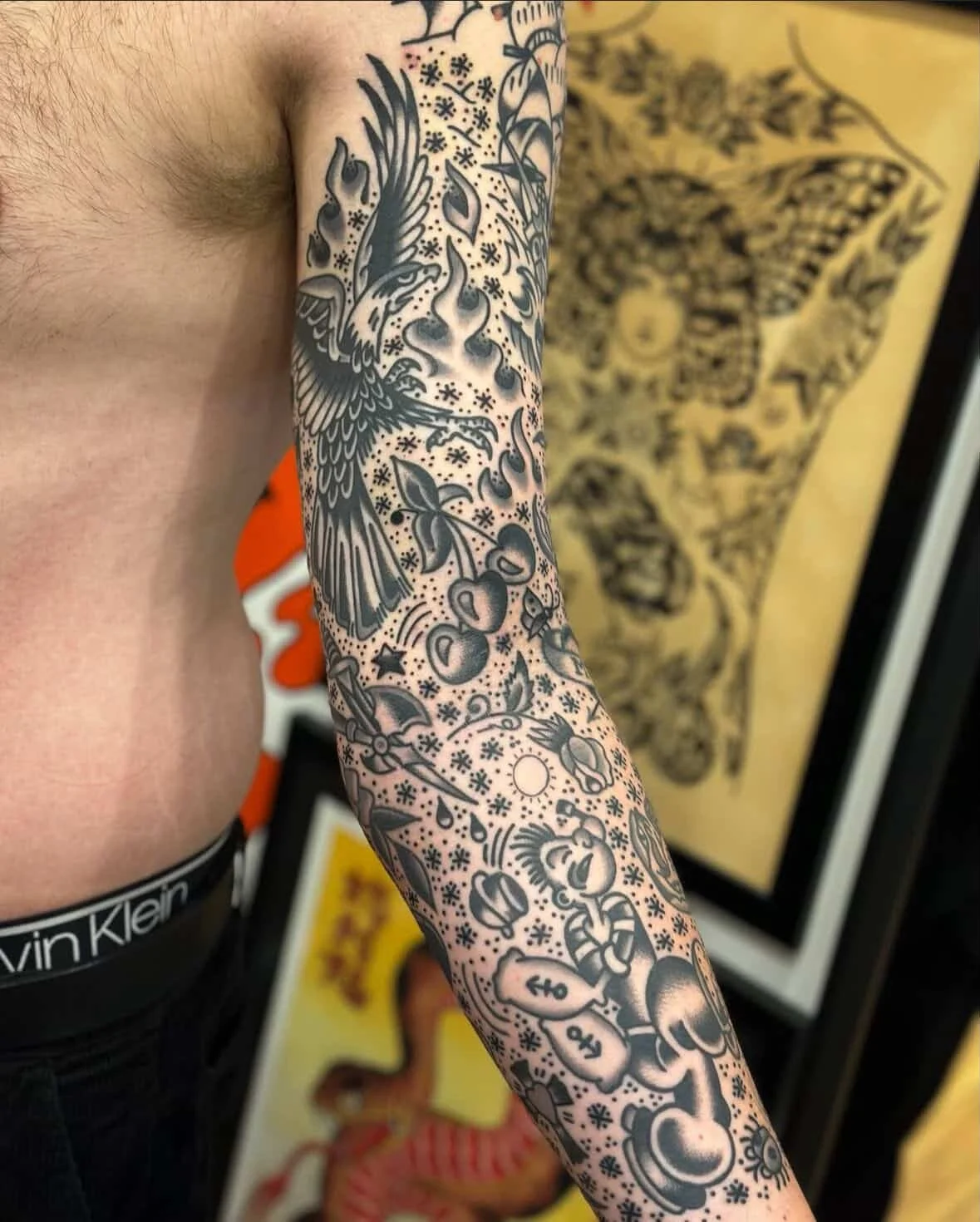 Tattoo sleeve on person's arm featuring various images including a bird, cherries, a sun, a skull, and other decorative elements in black ink.