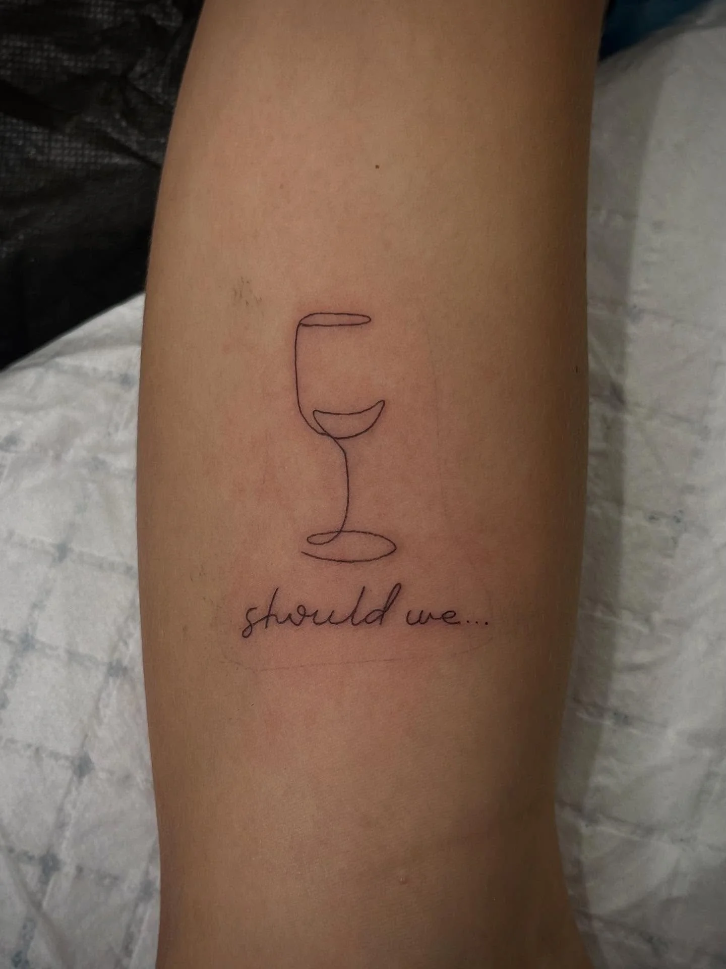 Tattoo of a line drawing of a wine glass with the phrase "should we..." below it.