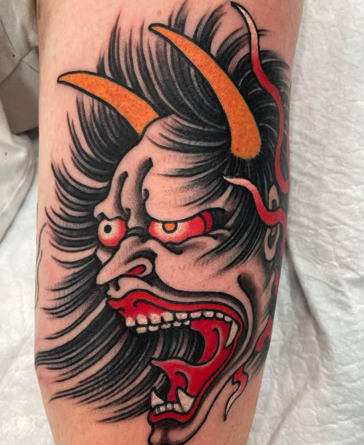 Tattoo of a traditional Japanese demon mask with exaggerated facial features, sharp teeth, red tongue, yellow horns, and flowing black hair.