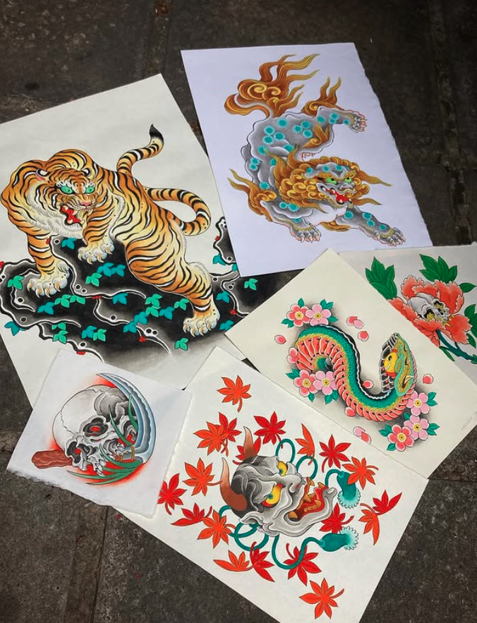 A collection of colorful traditional Asian tattoo-style drawings on paper laid out on a concrete surface. The drawings include a tiger, a dragon, a snake, a skull, and a demon mask surrounded by flowers and leaves.