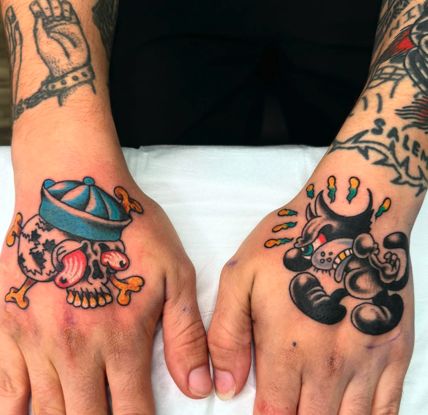 Hand tattoos of a skull with a blue hat and orange bones on the left hand, and a cartoon character with a necktie and a cigar on the right hand.