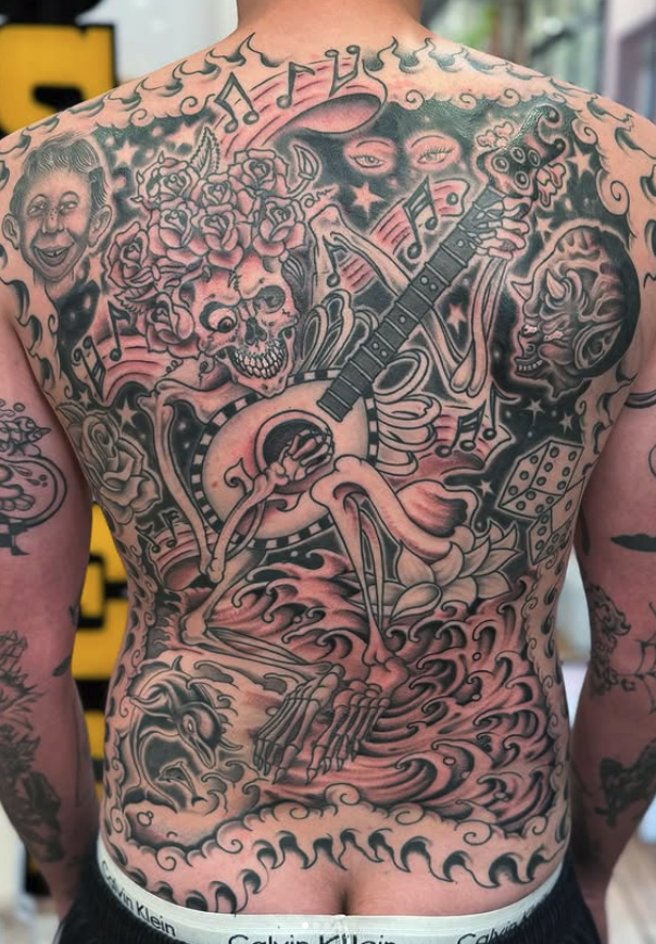 A detailed black and gray tattoo covering the person's chest and torso. It includes a smiling face, a skull with flowers on top, musical notes, a guitar, a dice, a woman's face, waves, clouds, a dragon, and skeletal hands, creating a surreal and arti