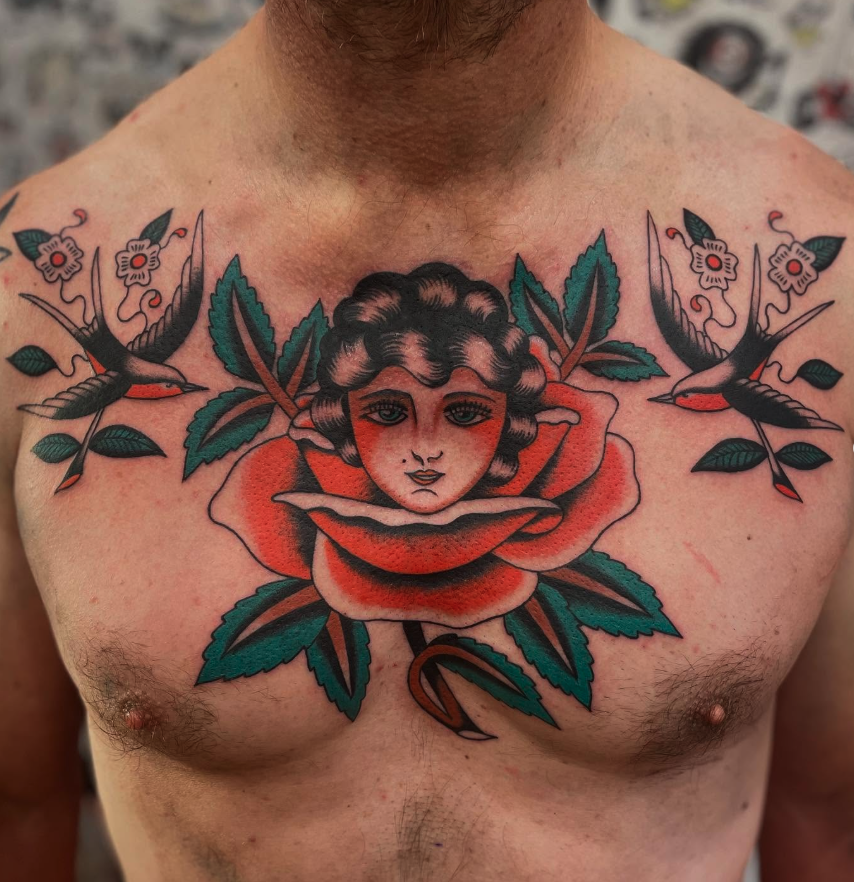 Tattoo of a woman's face with curly hair surrounded by a large red and white rose, green leaves, and two swallows flying on either side, on a man's chest.