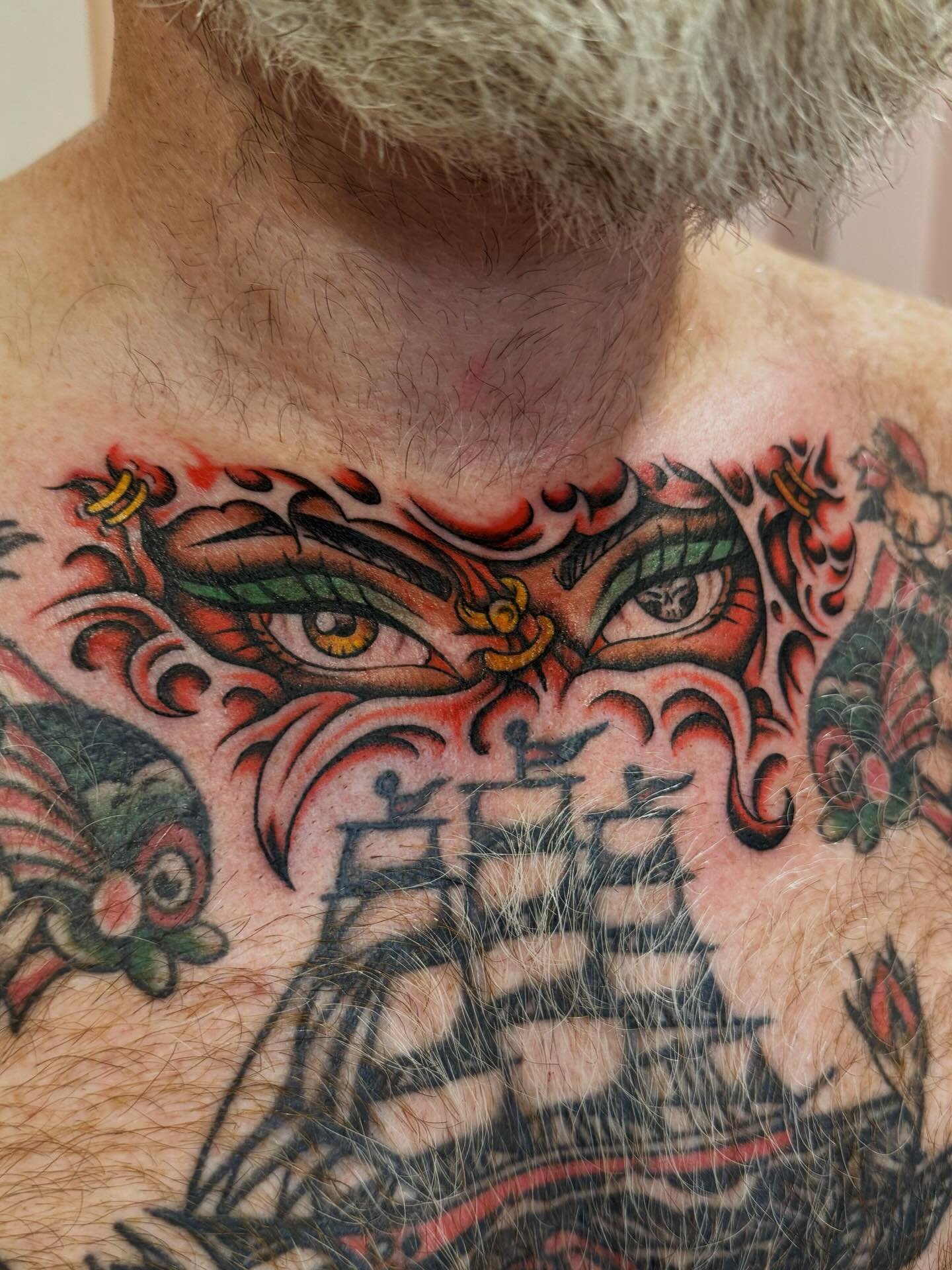 Close-up of a tattooed chest and neck, featuring a colorful design of a goddess or mythological figure with prominent eyes, green eyebrows, and ornate details.