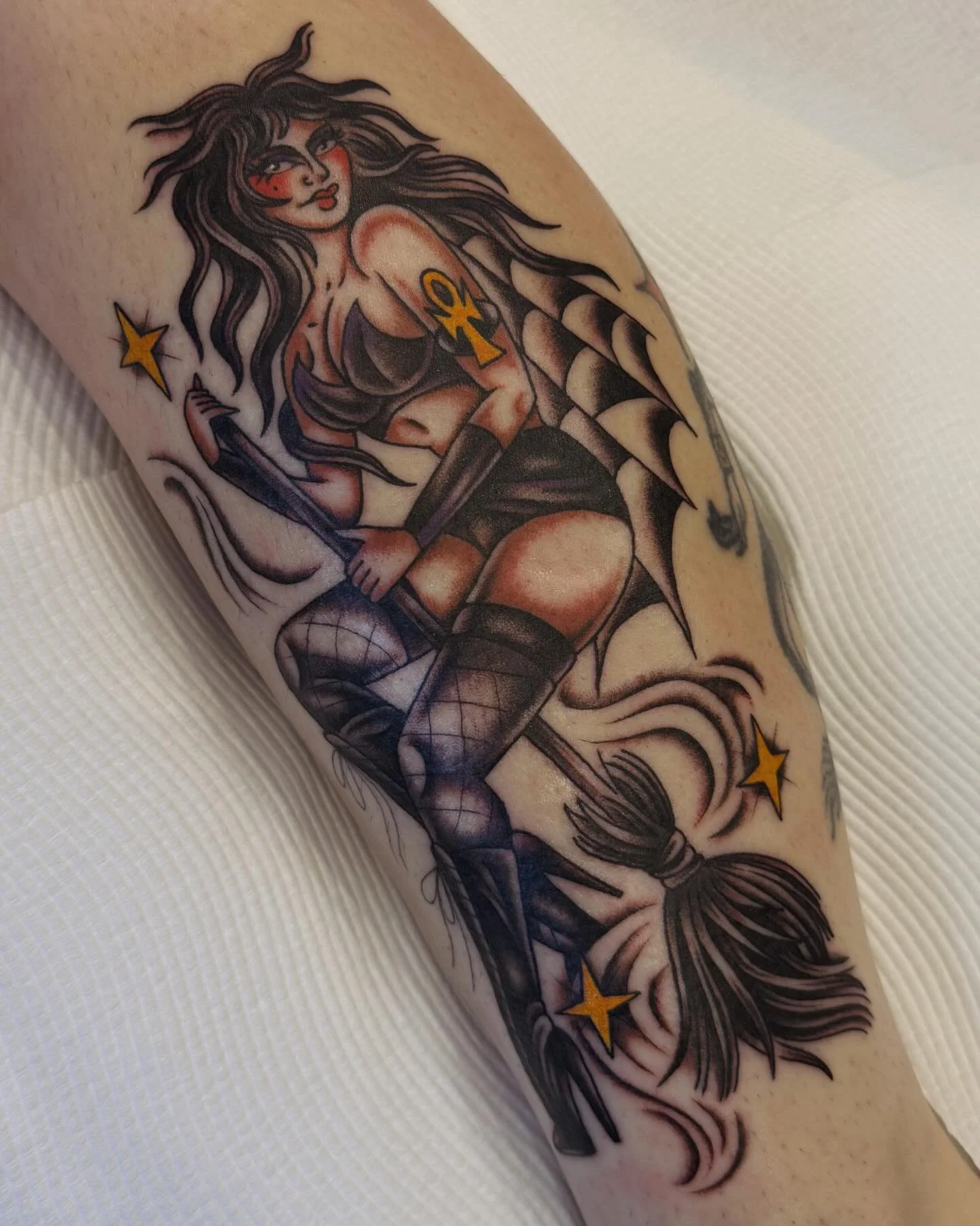 Tattoo of a woman with long, wavy hair, wearing black lingerie, thigh-high stockings, and holding a broom, surrounded by stars and bats.