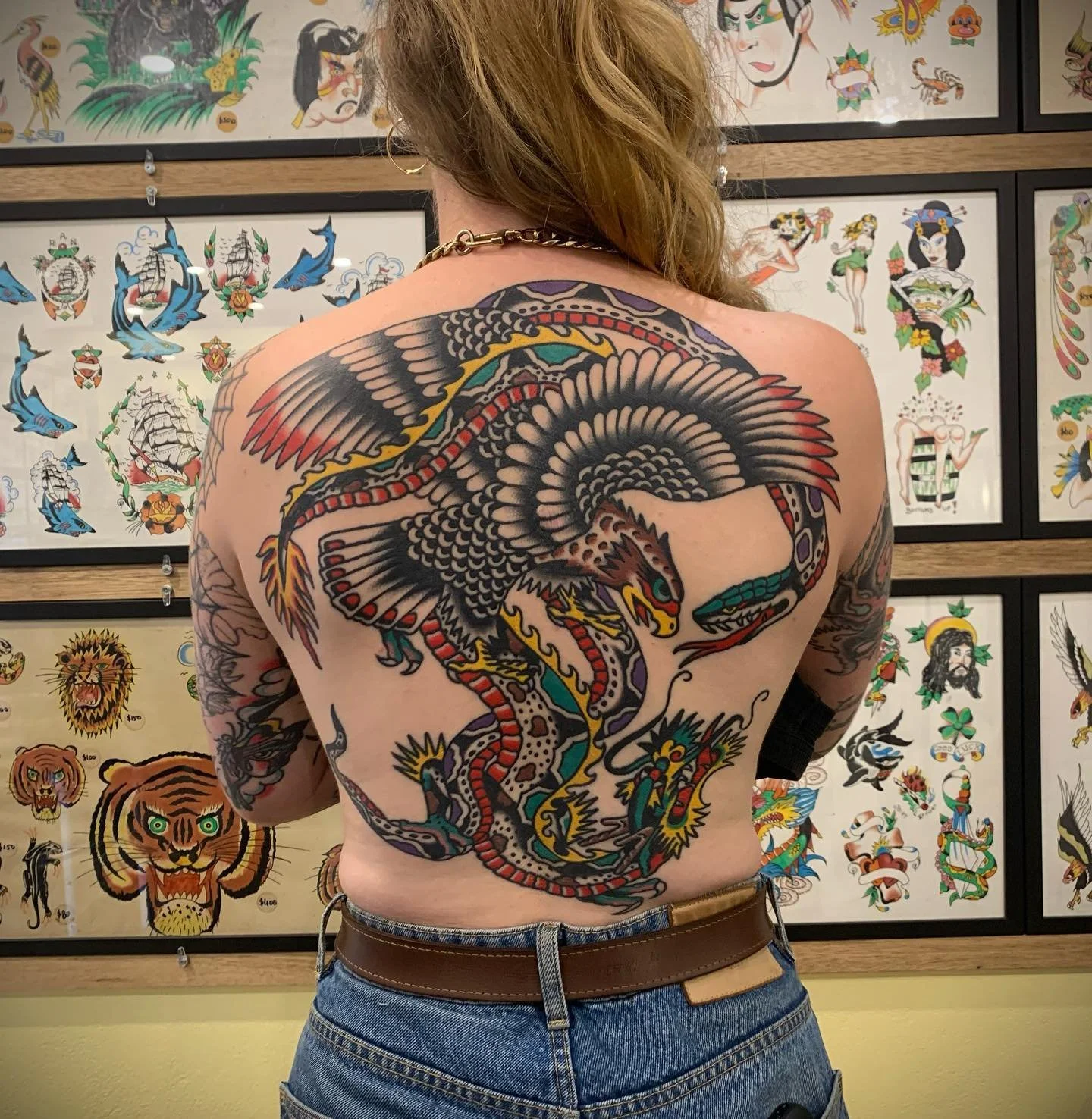 A person with a large, colorful dragon tattoo covering their back, surrounded by artwork and traditional tattoo flash designs on the wall.