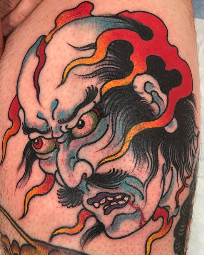 Tattoo of a fierce, stylized face with intense eyes, prominent mustache, and expressive features, with swirling red, yellow, and blue accents.