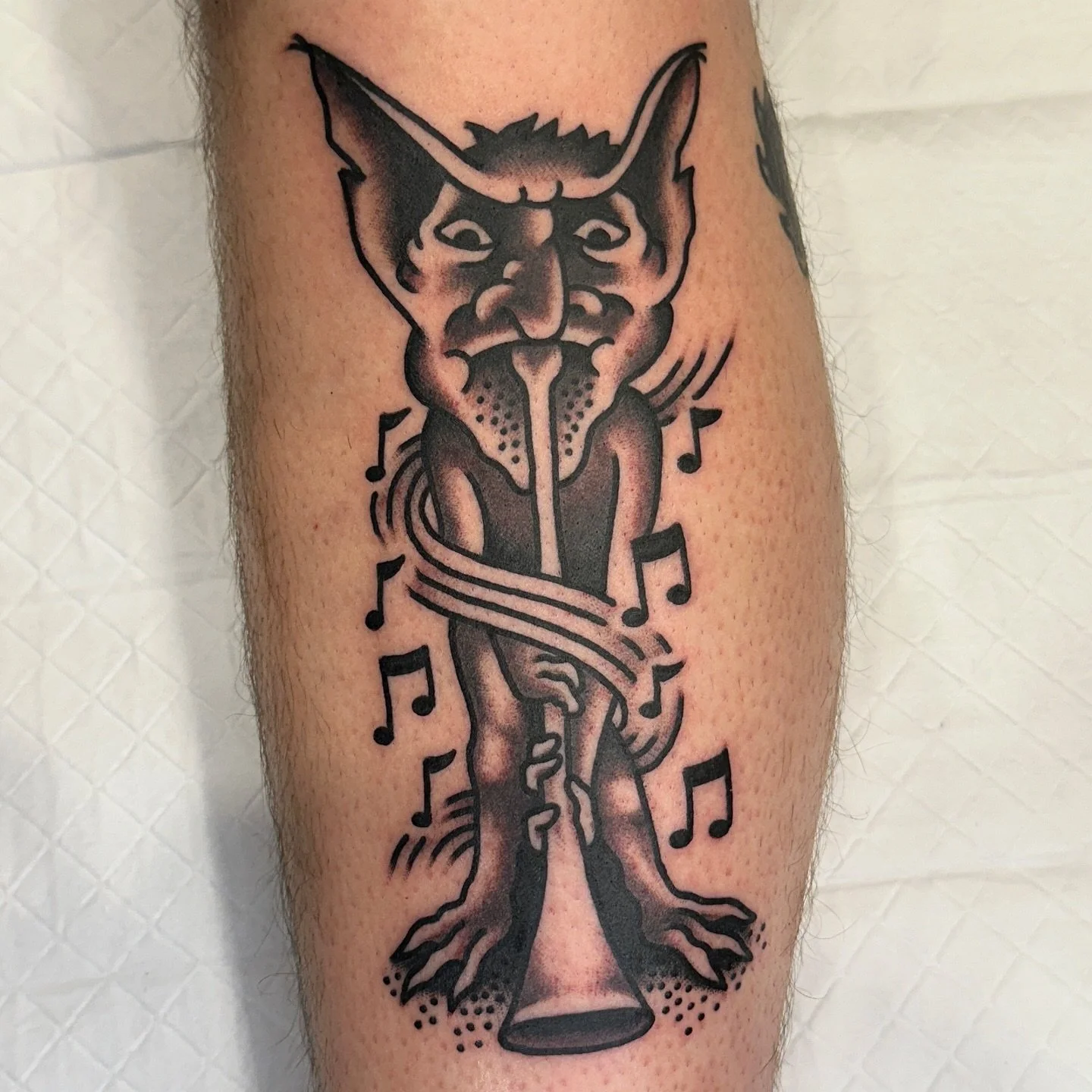 Tattoo of a devil-like creature with large ears, horns, and a long tongue, playing a musical instrument shaped like a big bell, surrounded by musical notes.