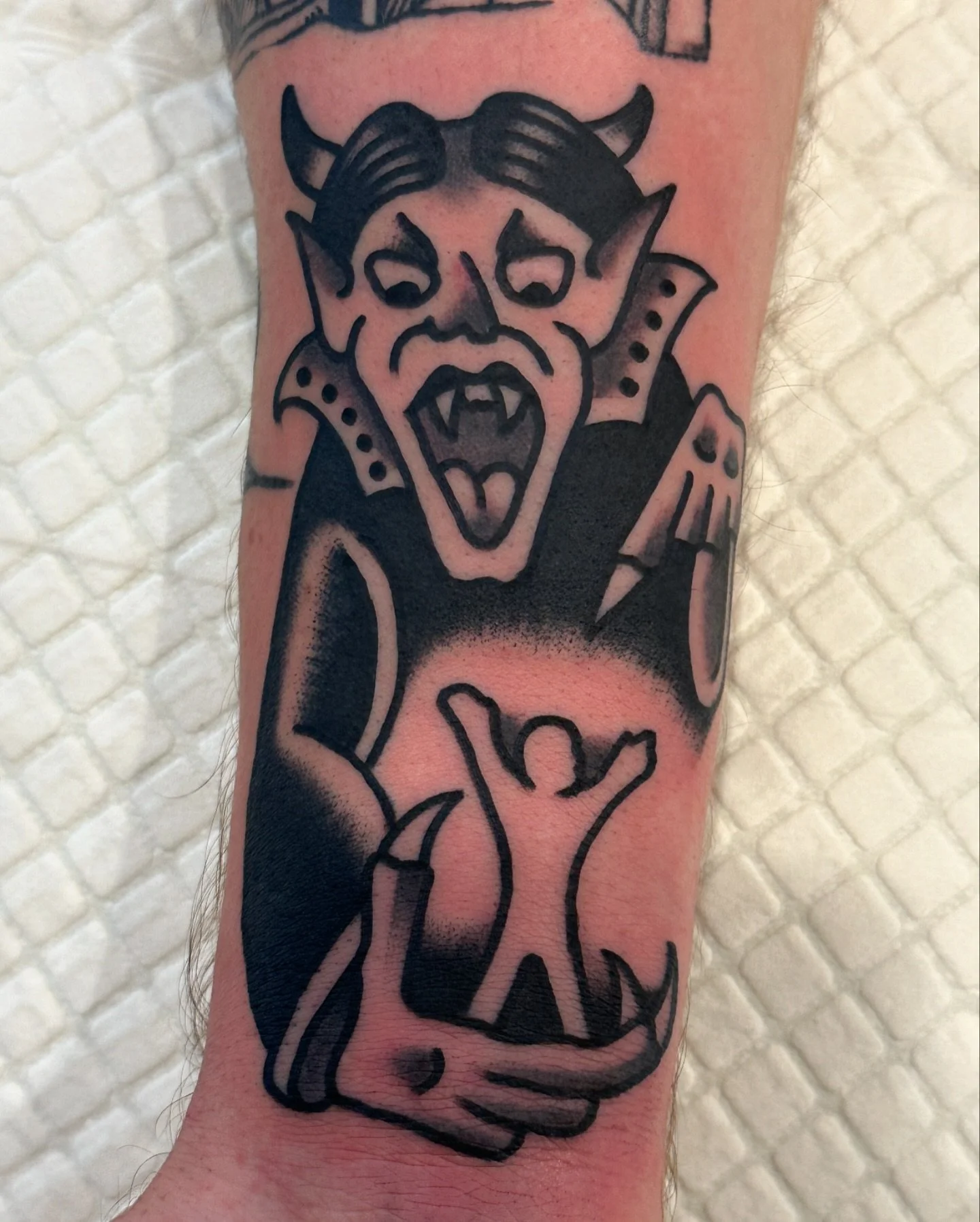 Tattoo of a demon-like figure with horns, sharp teeth, and pointy ears, holding a devil's pitchfork, on a person's arm.