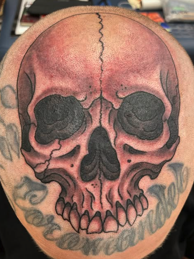 A tattoo of a realistic skull on a person's shoulder with detailed shading and black accents, surrounded by other tattoos.