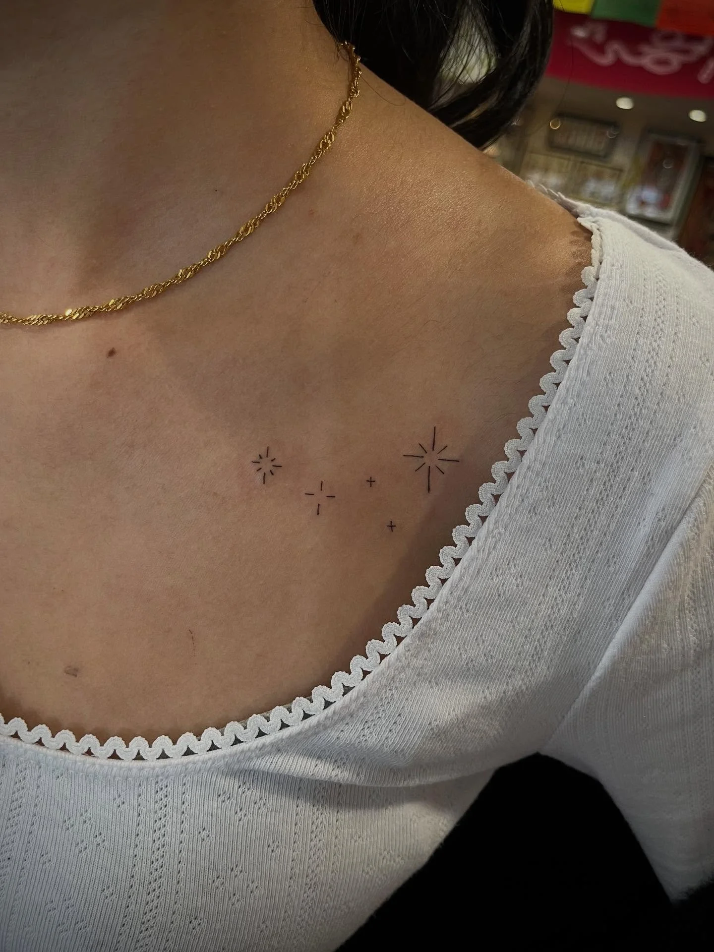 Close-up of a woman's shoulder and collarbone area with small tattoos of stars and plus signs, wearing a white top with a scalloped neckline and a gold chain necklace.