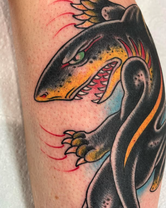 Colorful tattoo of a mythical creature with a dragon-like face, sharp teeth, green eye, yellow and black coloring, claws, and a black body, on a person’s arm.