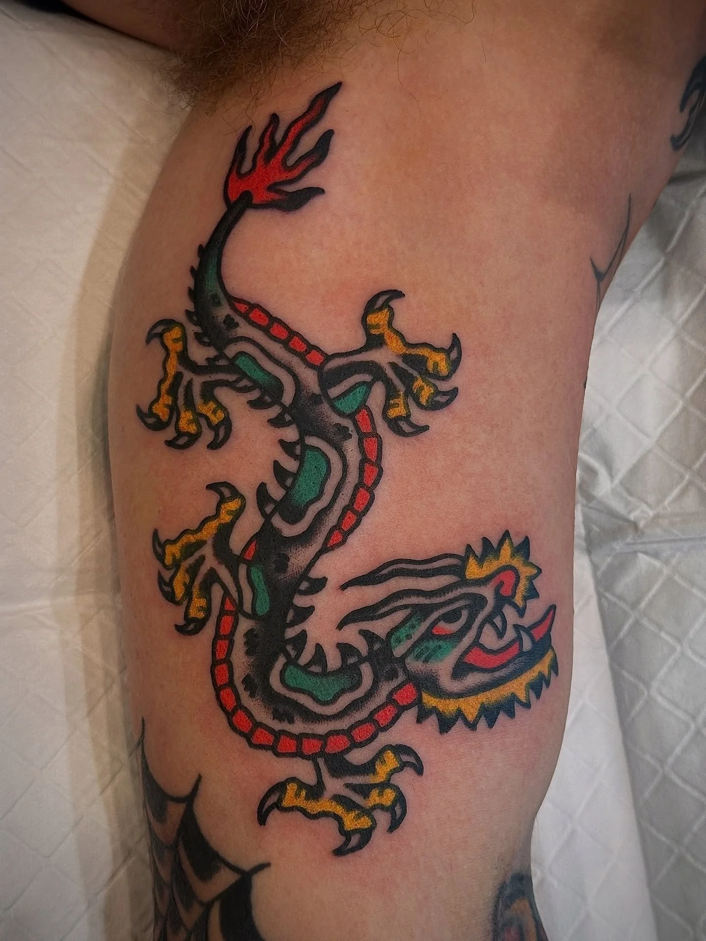Tattoo of a colorful dragon with black outlines, red, green, yellow, and orange accents on person's upper arm.