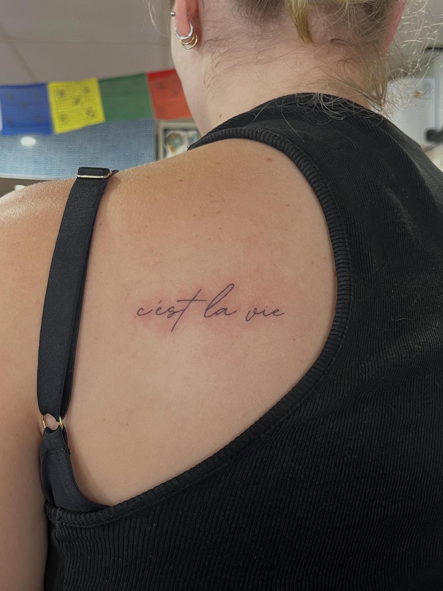 A close-up of a woman's shoulder and upper back, showing a tattoo with the French phrase 'c'est la vie' in thin cursive script on her shoulder blade, with part of her ear and earrings also visible.