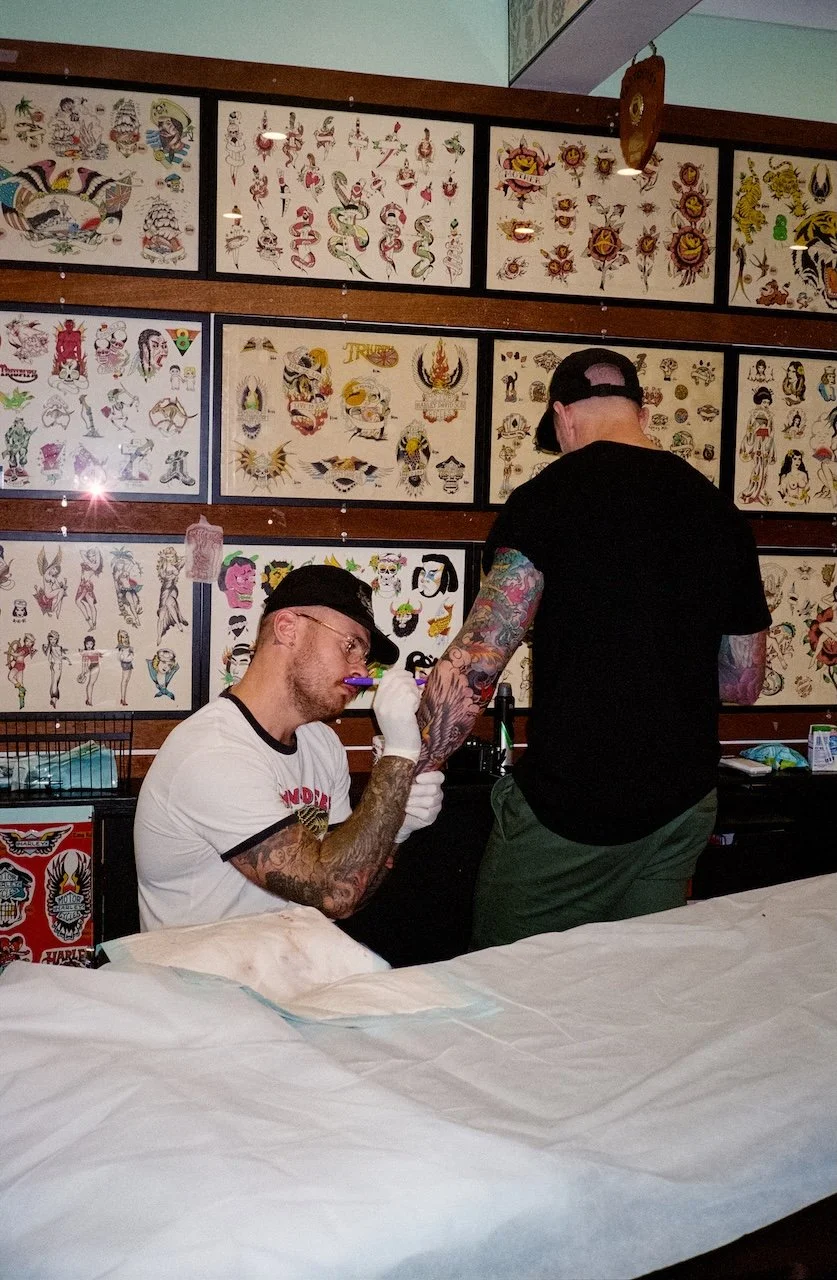 A man getting a tattoo on his arm at a tattoo studio, with tattoo flash art displayed on the wall behind him.