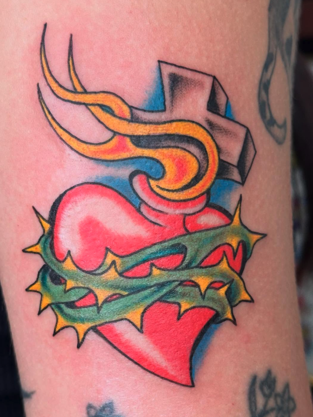 Tattoo of a broken red heart with a yellow and green thorny vine wrapped around it, with a black and gray cross behind the heart.