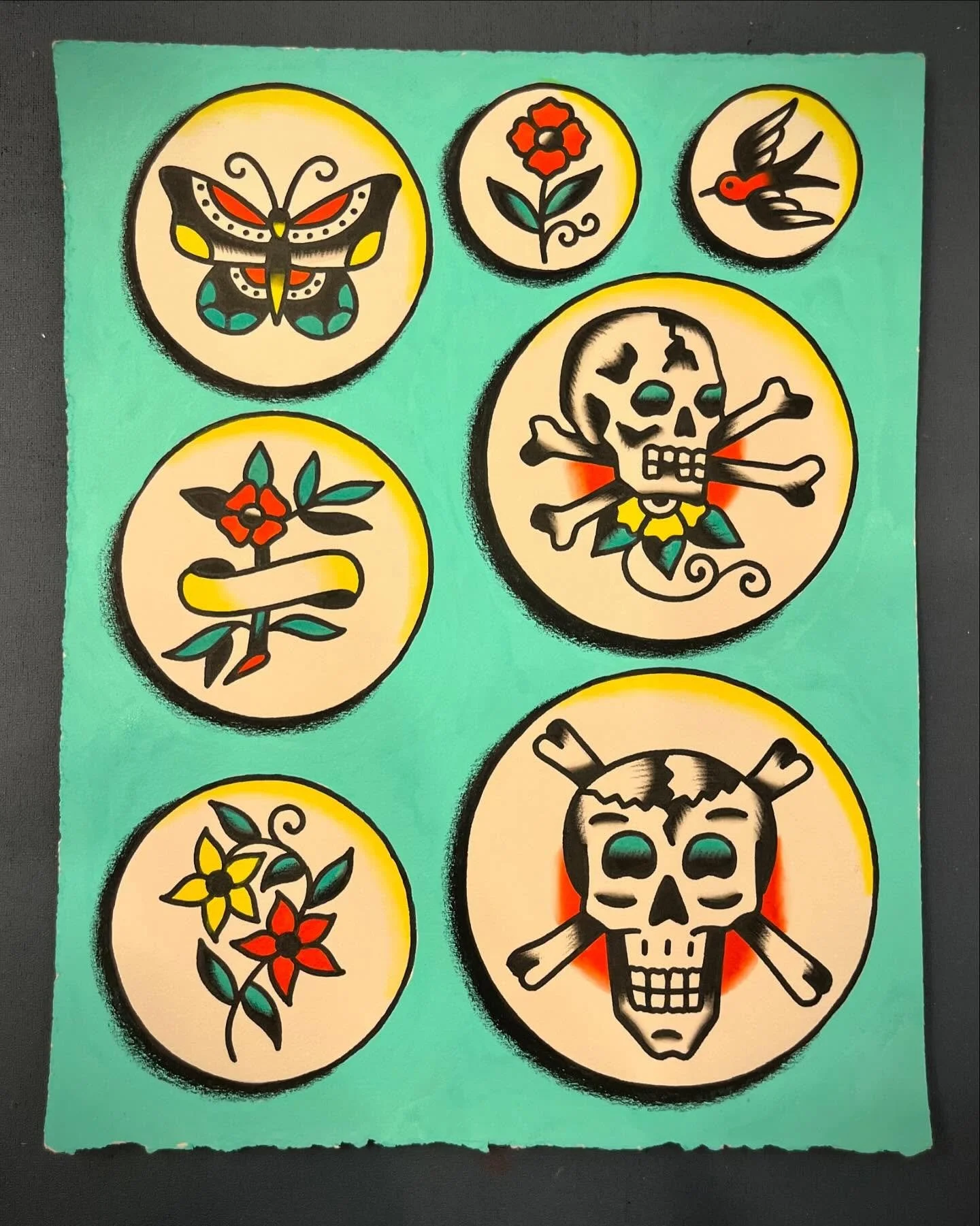 Tattoo flash sheet featuring colorful traditional-style skulls, flowers, butterflies, and birds with bold black outlines on a turquoise background.