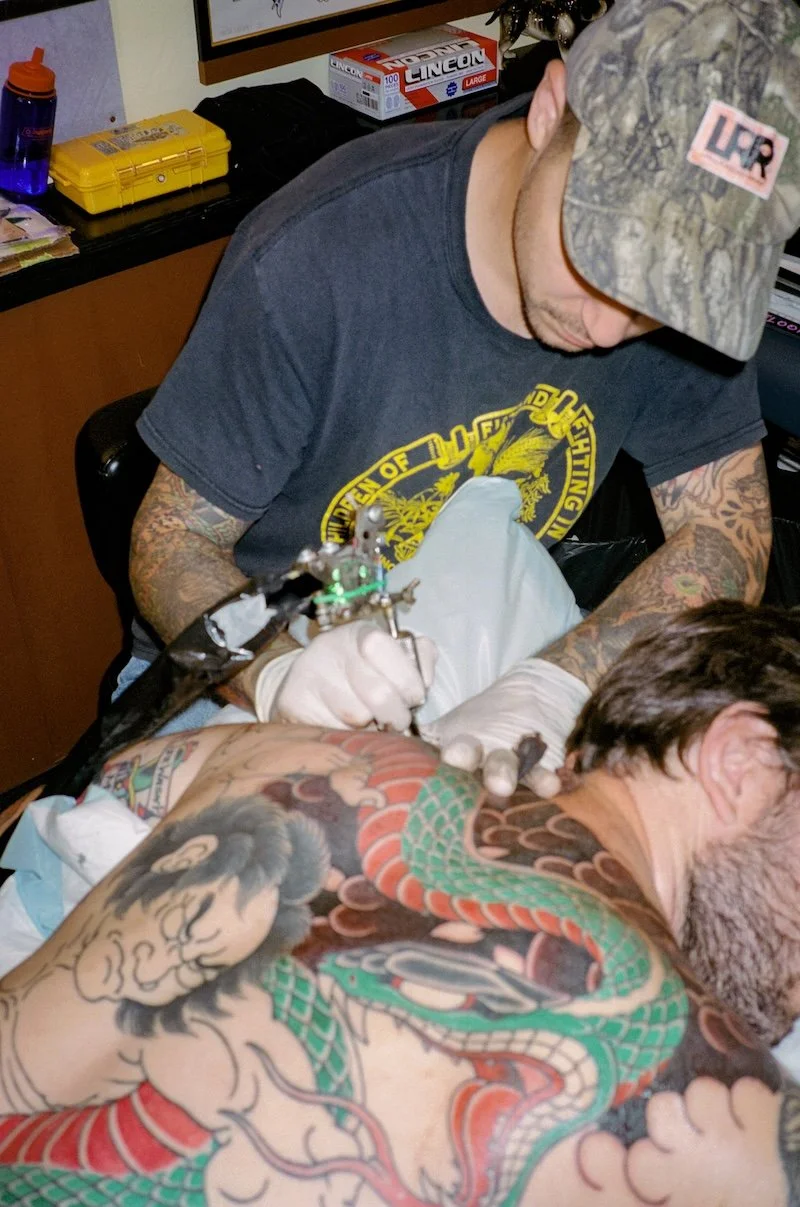 A tattoo artist wearing white gloves, a black t-shirt with yellow graphics, and a camouflage cap is tattooing a colorful dragon tattoo on a client’s arm. The client is lying down, and the scene takes place in a tattoo studio with various supplies and tools visible in the background.