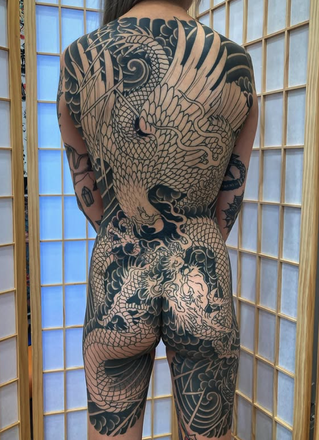 A person with extensive black and white traditional Japanese-style dragon tattoos covering their back, buttocks, and legs, standing in front of a wooden folding screen.