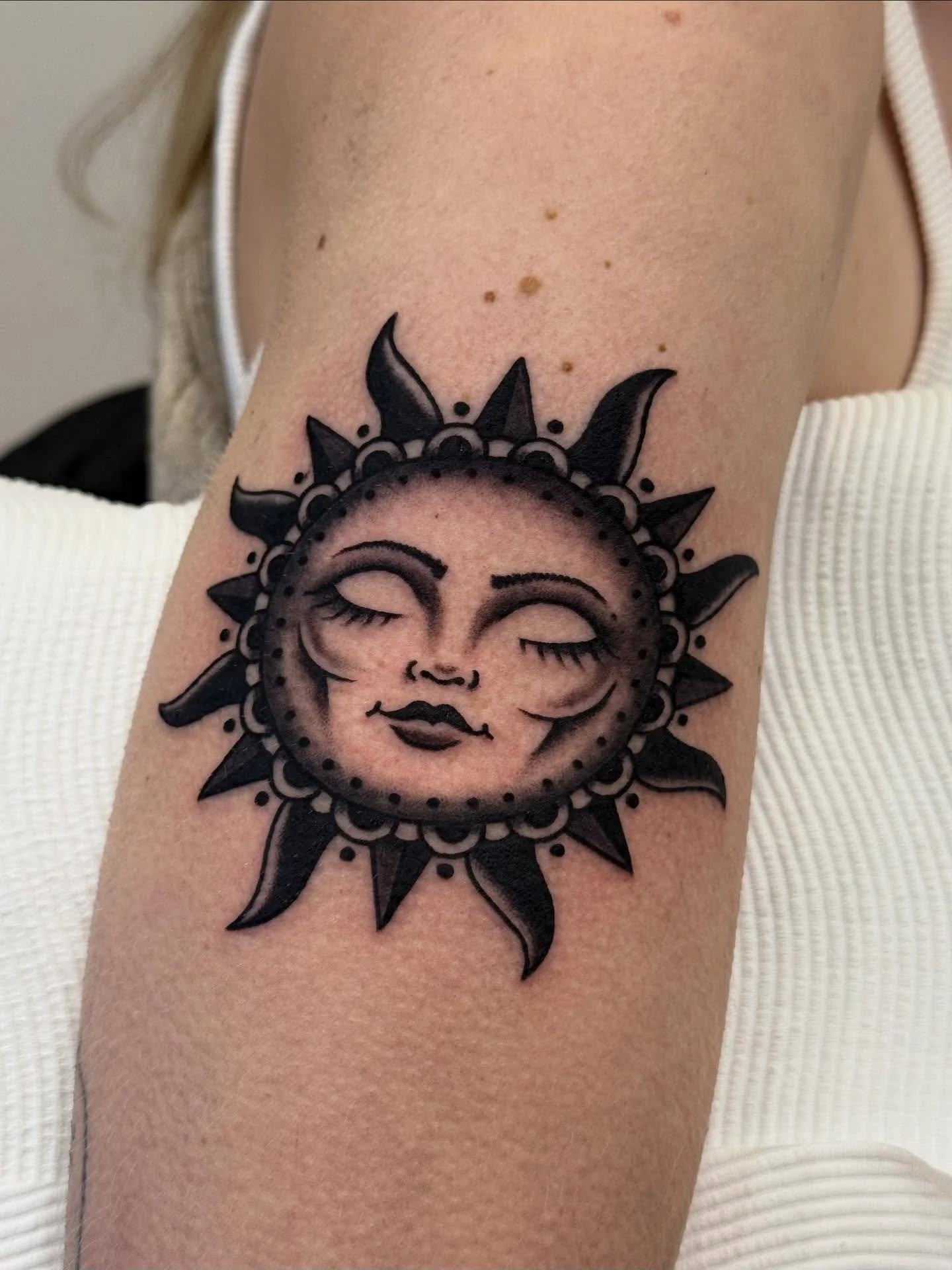 Black sun tattoo with a woman's face in the center, closed eyes, lips, and stylized hair, on a person's upper arm.