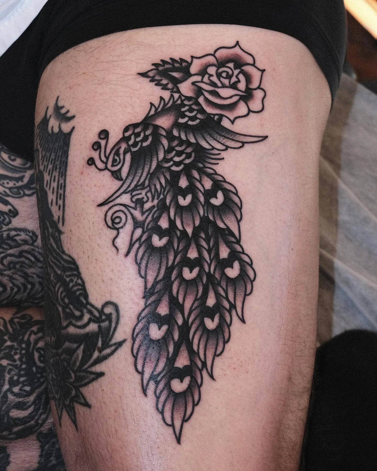 A black tattoo of a bird with a rose in its beak and a tail made of heart-shaped leaves on a person's upper thigh.
