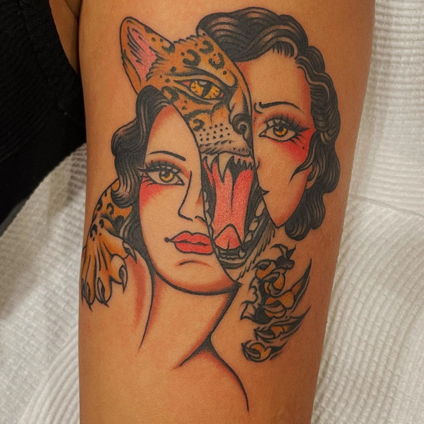 Tattoo of a woman's face with dark curly hair, bold features, and red blush, combined with a jaguar's face with golden eyes and open mouth showing sharp teeth, merging in the center.