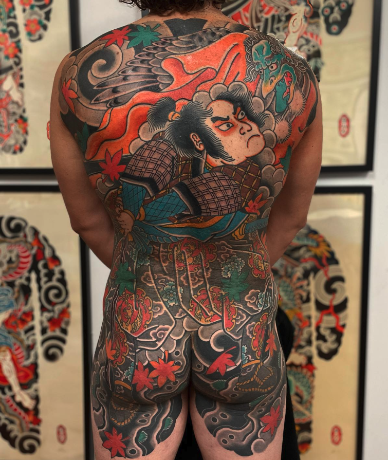 Full back and upper legs tattoo of a traditional Japanese woodworking figure and a dragon, with colorful maple leaves, clouds, and waves.